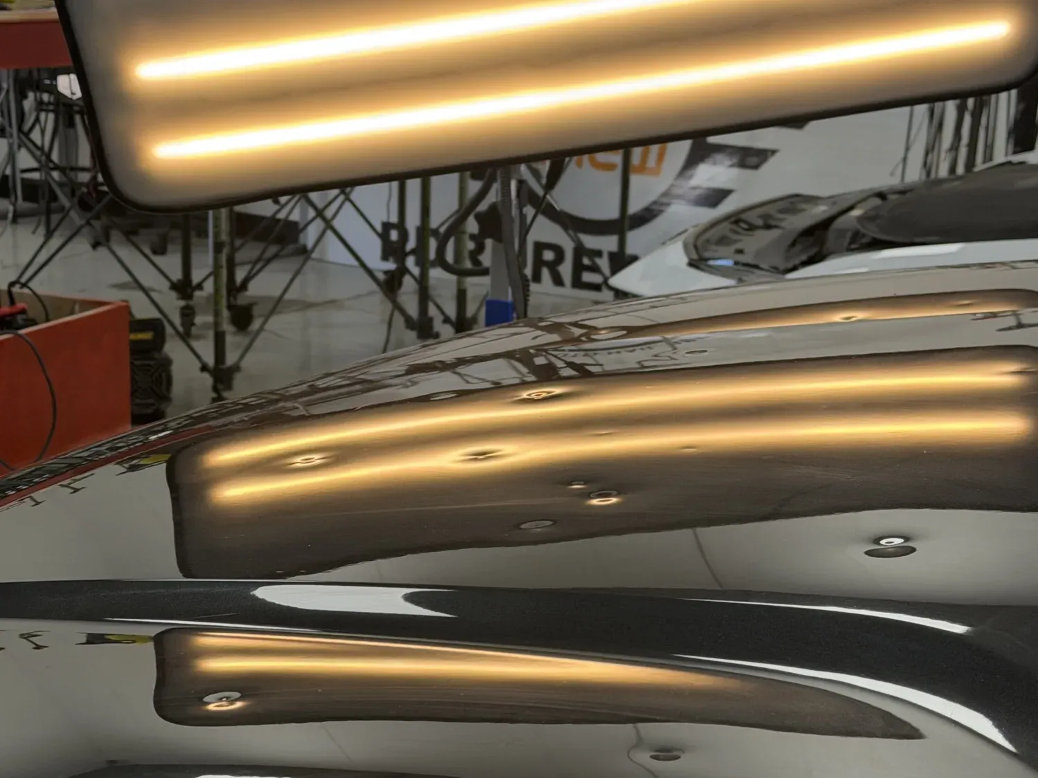 Black hood under a PDR line board, hail dents before and straight reflections after the repair — before