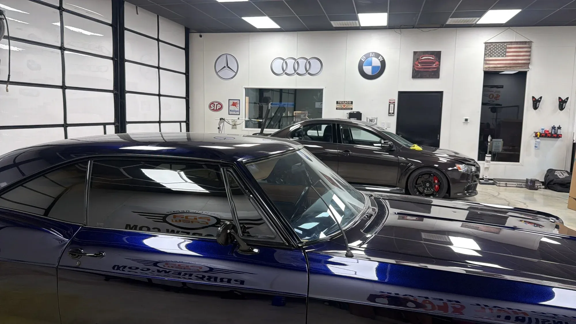 Inside the PDR Crew shop: a classic blue Chevelle with the PDR Crew logo, a black Lancer Evolution and Mercedes, Audi and BMW logos on the wall