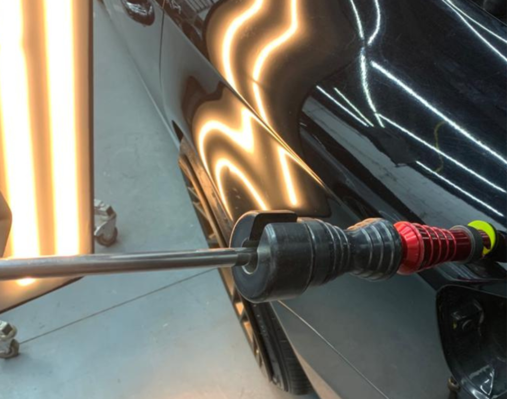 A close-up of a paintless dent repair tool in use on a black car. The tool is attached to the car's surface, and bright lights are illuminating the area, reflecting off the car's shiny surface. The setup highlights the precision and technique used in the dent repair process.