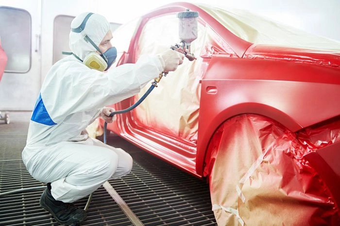 A technician in a full protective suit, including a respirator and goggles, is painting a red car in a spray booth. The car is carefully masked off with plastic and tape to protect areas not being painted. The technician is crouching and using a spray gun to apply an even coat of paint to the car's surface. The bright, controlled environment of the spray booth ensures a high-quality finish. The technician's attention to detail and precision in applying the paint are evident as he focuses on achieving a smooth and uniform application.