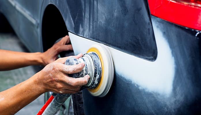 A technician is using an orbital sander to smooth the surface of a car before painting. The car's rear quarter panel has been sanded down, preparing it for the next stages of repair and painting. The technician's hands are covered in sanding dust, emphasizing the hands-on nature of the work. The image highlights the attention to detail and precision required in the car repair and refinishing process. The car's surface shows signs of preparation, ready for the application of primer and paint.