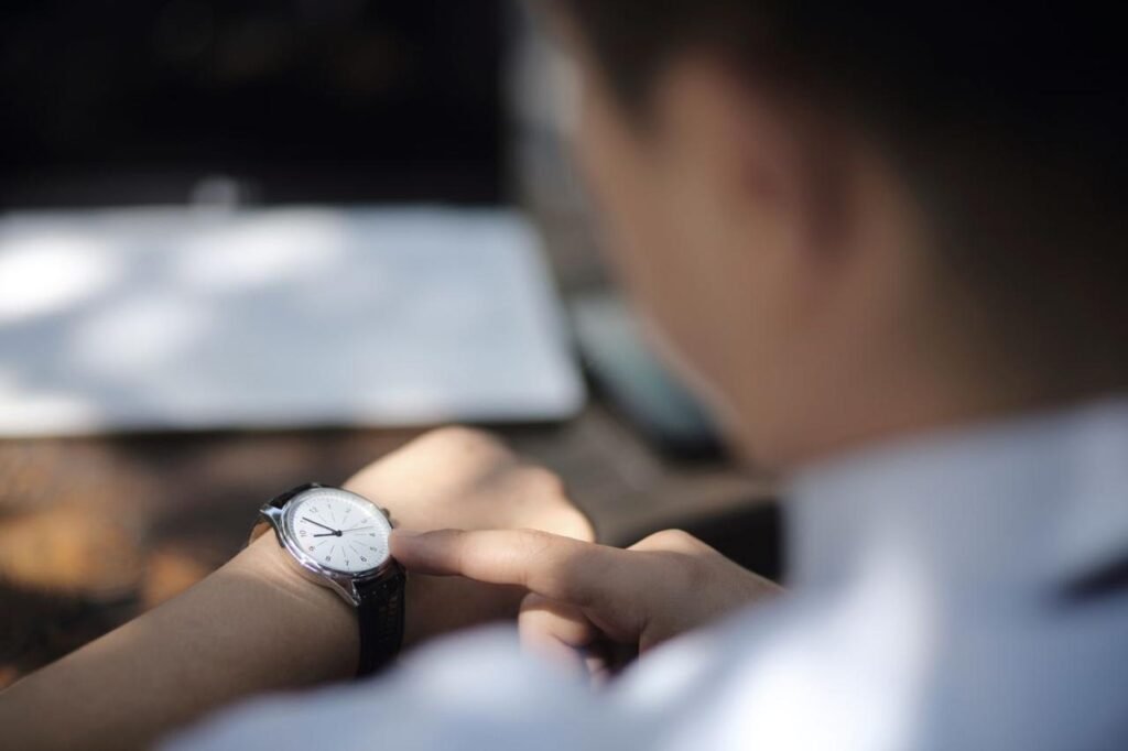 A close-up of a person looking at their wristwatch. The watch has a white face with black hands and a black leather strap. The person's finger is pointing at the watch. The background is blurred, showing what appears to be a laptop and a phone on a table.