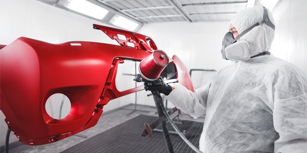 A technician in a full protective suit, including a respirator, is painting a red car bumper in a spray booth. The bumper is mounted on a stand, allowing the technician to apply an even coat of paint to all surfaces. The well-lit, controlled environment of the spray booth ensures a high-quality finish. The technician's attention to detail and precision in applying the paint are evident as he focuses on achieving a smooth and uniform application.