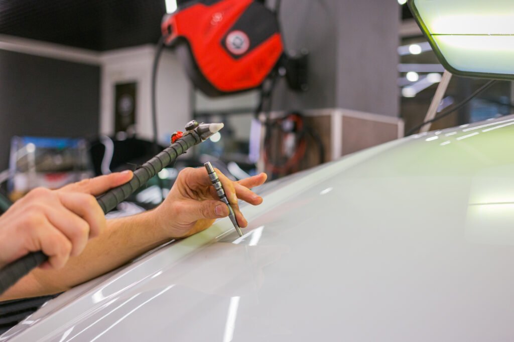 A close-up of a person performing paintless dent repair on a car. The technician's hands are using specialized tools to fix a dent on the car's surface. In the background, a red tool bag and other equipment are visible in a well-lit garage setting.