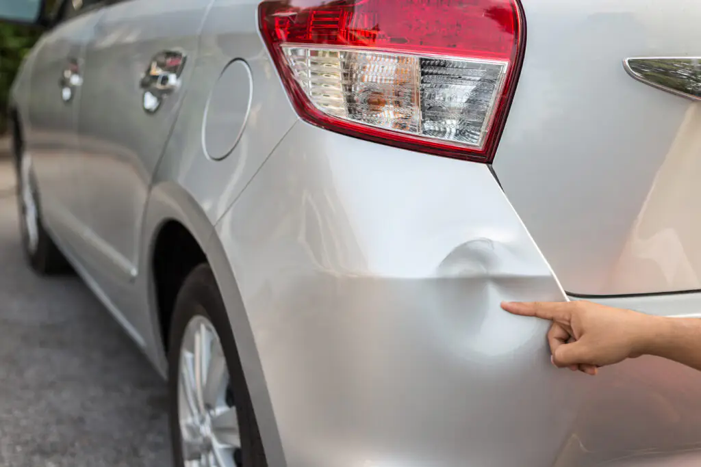 A close-up of a silver car with a noticeable dent near the rear taillight. A person's hand is pointing at the dent, emphasizing the damage. The car is parked on a street, and the taillight, fuel door, and rear wheel are visible in the image. The dent appears to be a common type of damage that could be repaired using paintless dent repair techniques.